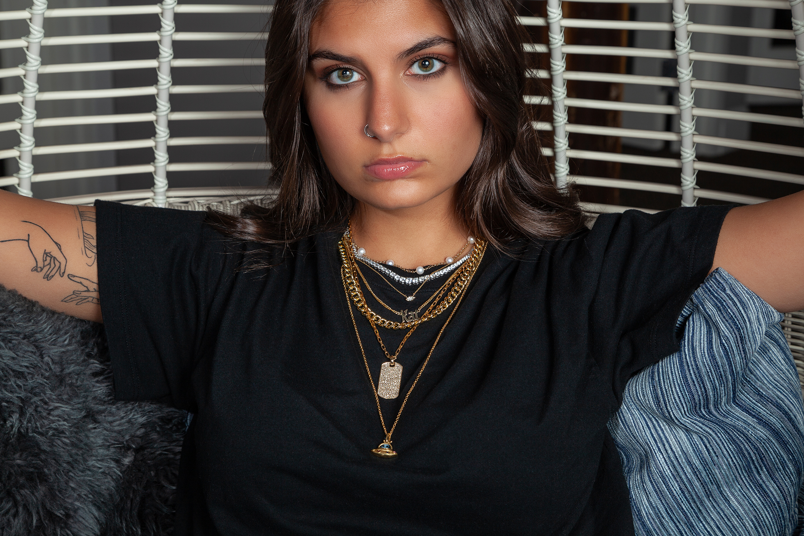 model with striking eyes and dark hair wearing a black tshirt and layered gold chains stares directly into the camera